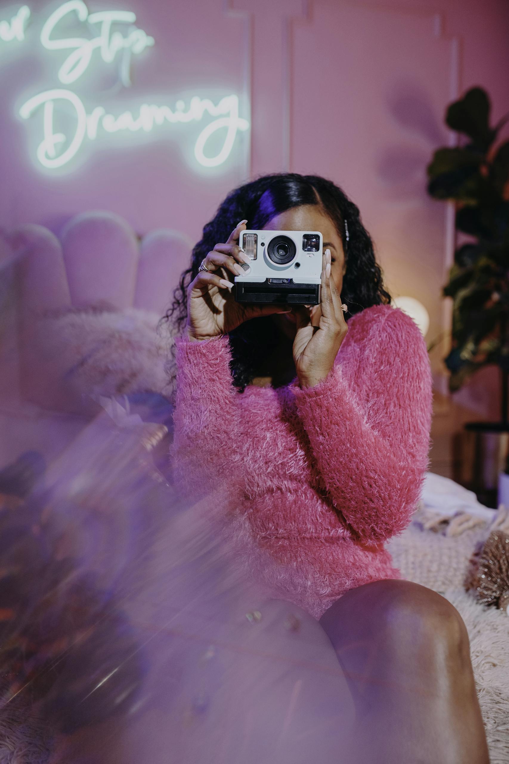 A fashionable woman holds a vintage camera in a cozy, neon-lit setting.